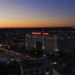 Aerial view of a cityscape at dusk with the sky glowing orange. A large building with illuminated “Montgomery Plaza” signs is visible, surrounded by city lights and busy streets filled with cars.