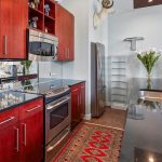 Modern kitchen with shiny black countertops, red wooden cabinets, stainless steel appliances, a red rug, decorative vases, and a mounted deer head on the wall. A bouquet of white flowers sits near a fridge.