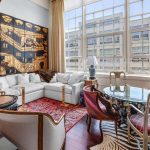 A stylish living room with a white sectional sofa, eclectic chairs around a glass table, a vibrant rug, zebra-patterned rug, large windows with city views, and a large black and gold mural on one wall.