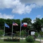 Three flags—the Oklahoma state flag, the U.S. flag, and the Texas state flag—fly on flagpoles in front of a gated park area with green trees and a partly cloudy sky in the background.