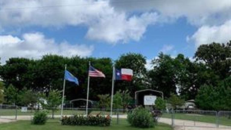 Three flags—the Oklahoma state flag, the U.S. flag, and the Texas state flag—fly on flagpoles in front of a gated park area with green trees and a partly cloudy sky in the background.