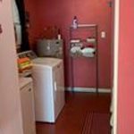 A narrow laundry room with red walls features a washer, dryer, folded towels on a metal rack, and a small rug on the floor. A ceiling fan is visible above.