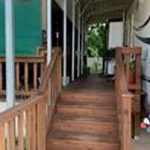 A wooden staircase with railings leads up to a covered porch area with a green screen on the left and potted plants in the background. The porch has a metal roof and is surrounded by greenery.