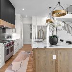 Modern kitchen with white cabinets, a wood-accent island, gold fixtures, stainless steel appliances, a black backsplash, and pendant lights. A vase with white orchids sits on the island; stairs are visible in the background.