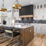 Modern kitchen with white cabinets, a large island with seating for three, gold and black accents, wood flooring, pendant lights, built-in appliances, and a vase with white flowers on the island.