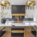 Modern kitchen with a large white island featuring black and gold accents, three black and gold chairs, a potted white orchid centerpiece, white cabinetry, and gold light fixtures above.