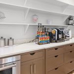 Modern kitchen with brown cabinets, white countertops, metal canisters, coffee machine, flavored syrup bottles, mixer, and glass jars on open shelves against a white wall.
