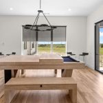 A modern game room with a light wood pool table and bench, large windows with a scenic outdoor view, sliding glass doors, and a black sectional sofa in the background.