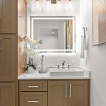 A modern bathroom with light brown cabinets, a white square sink, a wall-mounted mirror with LED lights, and a vase of white flowers on the countertop. Neutral decor and clean lines create a contemporary look.