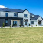 A modern two-story house with white and dark gray exterior, large windows, a metal roof, and minimal landscaping, set on a spacious green lawn under a clear blue sky.