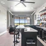 Modern home bar with black stools, a large wall-mounted TV, open shelves stocked with bottles, a sink, fridge, and wide windows showing a scenic outdoor view in the background.