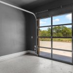 A room with dark gray walls, a light gray speckled floor, a large glass panel garage door, and a black door with a vault-style lock. Outside, there are trees, grass, and a blue sky with clouds.