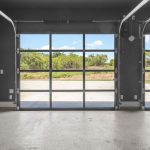 Interior view of an empty garage with a polished floor, dark walls, and two large glass-panel garage doors, revealing a driveway and green trees outside under a blue sky.