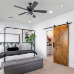 A modern bedroom with a four-poster bed, gray bench, wooden dresser, abstract art, a ceiling fan, a large plant, and a sliding barn door leading to a bathroom with white cabinets.