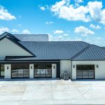 A modern, single-story white house with a dark metal roof, large garage doors, and glass windows stands under a bright blue sky with scattered clouds, surrounded by open, grassy fields.