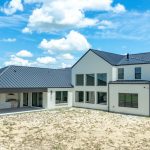 Modern two-story white house with black metal roof and large windows, featuring a covered patio area with outdoor furniture, set on a grassy and partially sandy yard under a blue sky with clouds.