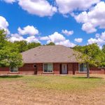 Single-story brick house with a gray shingled roof, surrounded by trees and a large dry lawn under a bright blue sky with scattered clouds.