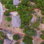 Aerial view of a house surrounded by trees, with a large irregularly-shaped lot outlined in red. The property is bordered by a curved road and neighboring houses, with grassy and wooded areas visible.