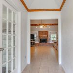 A tiled hallway with white walls, a glass-paneled door to the left, a hanging light fixture, and a view into a living room with a brick fireplace, wooden ceiling beams, and large windows.