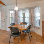 A cozy dining area with a wooden table, four black chairs, a hanging light above, floral curtains on three windows, and brick walls next to tiled flooring. A glass door leads outside. Flowers are on the table.