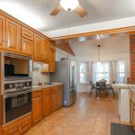 Spacious kitchen with wood cabinets, stainless steel appliances, brick accents, tiled countertops, and a ceiling fan. There's a dining area with windows in the background and a tile floor throughout.
