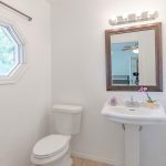 A bright bathroom with white walls features a pedestal sink, a toilet, a framed mirror, an octagonal window, and a small orchid on the sink. Light fixtures and a white shelving unit are also visible.