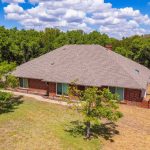 A single-story brick house with a large gray shingle roof sits in a spacious yard with patches of grass and a few trees, surrounded by a wooded area under a blue sky with scattered clouds.