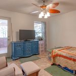 A cozy room with a white armchair, a blue dresser holding a TV, a bed with a colorful crocheted blanket, light tile flooring, and a ceiling fan. There’s a door and window letting in natural light.