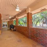 A covered brick porch with ceiling fans, outdoor furniture, and large rectangular openings looking out onto trees and a yard. The porch has a solid tan floor and red brick walls with white trim.