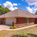 Single-story red brick house with a gray shingle roof, attached garage, and curved driveway. The yard has grass, small bushes, and mature trees under a partly cloudy blue sky.