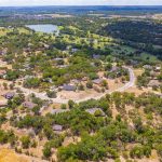 Aerial view of a suburban neighborhood surrounded by trees and greenery, with houses, a curving road, and a lake in the distance under a partly cloudy sky.