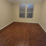 An empty room with wooden floors, beige walls, two windows with blinds, and a ceiling fan with a light fixture.