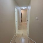 A well-lit hallway with beige tile floors and cream-colored walls, featuring several closed white doors and a light fixture on the ceiling. The hallway extends straight ahead into another room.