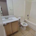A small bathroom with beige tile floor, a white toilet with the lid open, a bathtub with shower curtain rod, a sink with wood vanity, and a large wall mirror above the sink. The walls are light-colored.