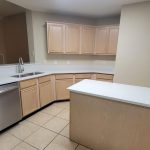 Modern kitchen with light wood cabinets, white countertops, a stainless steel dishwasher, under-mount sink, small island, and tiled floor. Neutral wall colors and recessed ceiling with bright overhead lighting.
