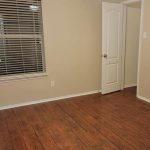 An empty room with beige walls, wooden flooring, a window with white blinds, a white door, and a ceiling fan with lights.