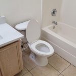 A clean bathroom with a white toilet, open lid, light wood vanity with sink and silver faucet, large mirror, and a white bathtub-shower combo; beige tiled floor and cream-colored walls.