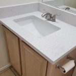 A small bathroom vanity with a speckled white countertop, built-in rectangular sink, silver faucet, light wood cabinets, a wall mirror, and a toilet paper holder mounted on the side.