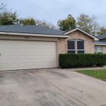 A single-story brick house with a two-car garage, a blue recycling bin near the driveway, trimmed bushes in front, and a cloudy sky overhead. Trees and neighboring houses are visible in the background.