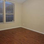 An empty room with light beige walls, two windows with white blinds, a ceiling fan with lights, and dark wood flooring. The room has a clean, minimal look and no furniture.