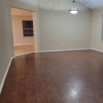 Empty room with wood flooring, beige walls, white trim, a decorative chandelier, and a window on the right. Tile flooring is visible in the foreground and through a wide doorway leading to another room.