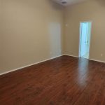 Empty room with dark wood floors, beige walls, a ceiling fan with lights, and a door leading to another room or bathroom. The ceiling is textured and the room has no furniture.