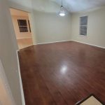 A spacious, empty room with dark wood floors, light beige walls, a window with blinds, a ceiling light fixture, and an open doorway leading to a tiled area.