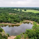 A small pond near County Road 419 in De Leon, TX 76444, surrounded by dense green trees and vegetation, with open grassy fields and cloudy skies visible in the background, viewed from above.