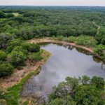 Aerial view of a small pond near County Road 419, De Leon TX 76444, surrounded by dense green trees and grassy patches, with a cloudy sky reflected on the water’s surface. The landscape extends into a forested area in the background.