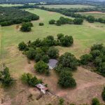 Aerial view of a rural landscape near County Road 419, De Leon TX, 76444, with open green fields, scattered trees, a house partially hidden by trees, and a small shed nearby. Wooded areas surround the land under a cloudy sky.
