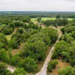 Aerial view of a rural landscape near County Road 419 in De Leon, TX 76444, with dense green trees, a winding dirt road, and open grassy areas under a cloudy sky.