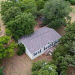 Aerial view of a single-story house with a gray roof on County Road 419, surrounded by large green trees and grass in the rural area of De Leon TX 76444.