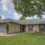 A single-story brick house with a gray shingle roof, two-car garage, large windows with black shutters, a well-kept lawn, and a mature tree in the front yard under a partly cloudy sky.