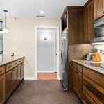Modern kitchen with granite countertops, wooden cabinets, stainless steel appliances, electric cooktop, and a microwave. The kitchen opens to a living area with a sofa and ceiling fan visible in the background.